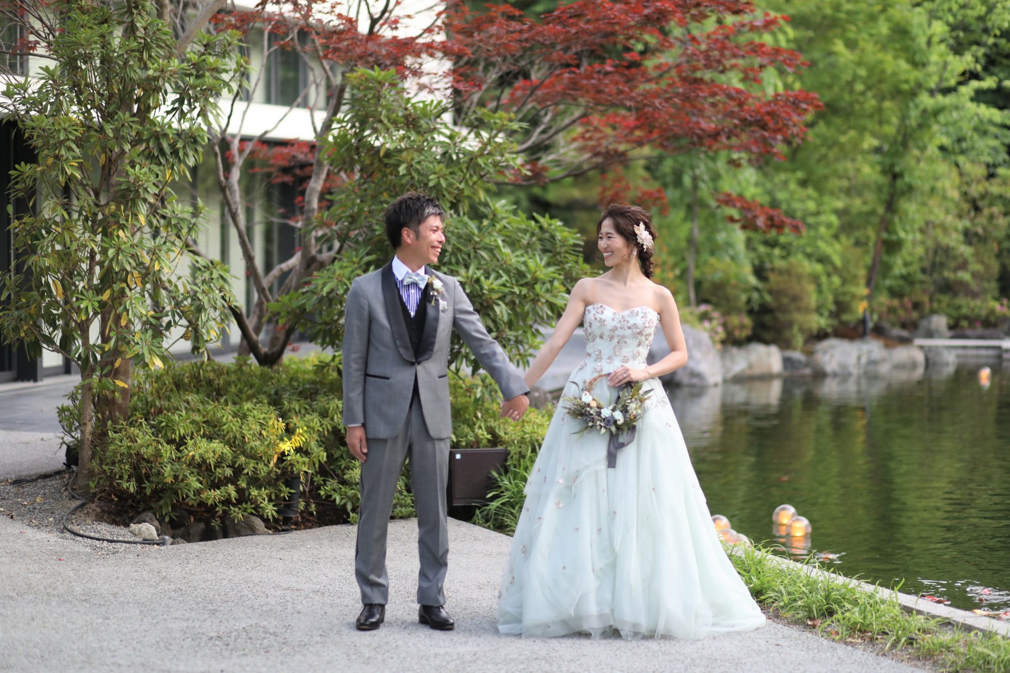 新緑の時期のザ・ガーデンオリエンタルオオサカでグレーのタキシードとモニークルイリエのカラードレスで手繋ぎショットされる新郎新婦様