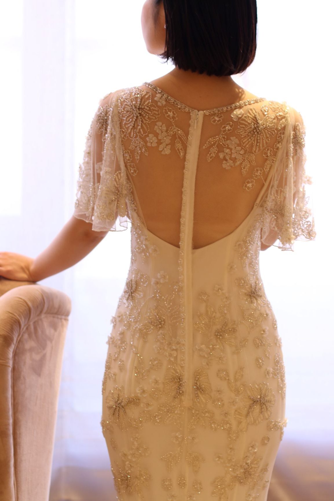 大きく空いたお背中に浮かび上がるようなフラワーモチーフのビーディングが、ゲストを魅了する圧巻のバックスタイルを印象付けるドレス前撮りや披露宴の衣装をお探しの方へご紹介したい袖付きのワンピース風ドレス