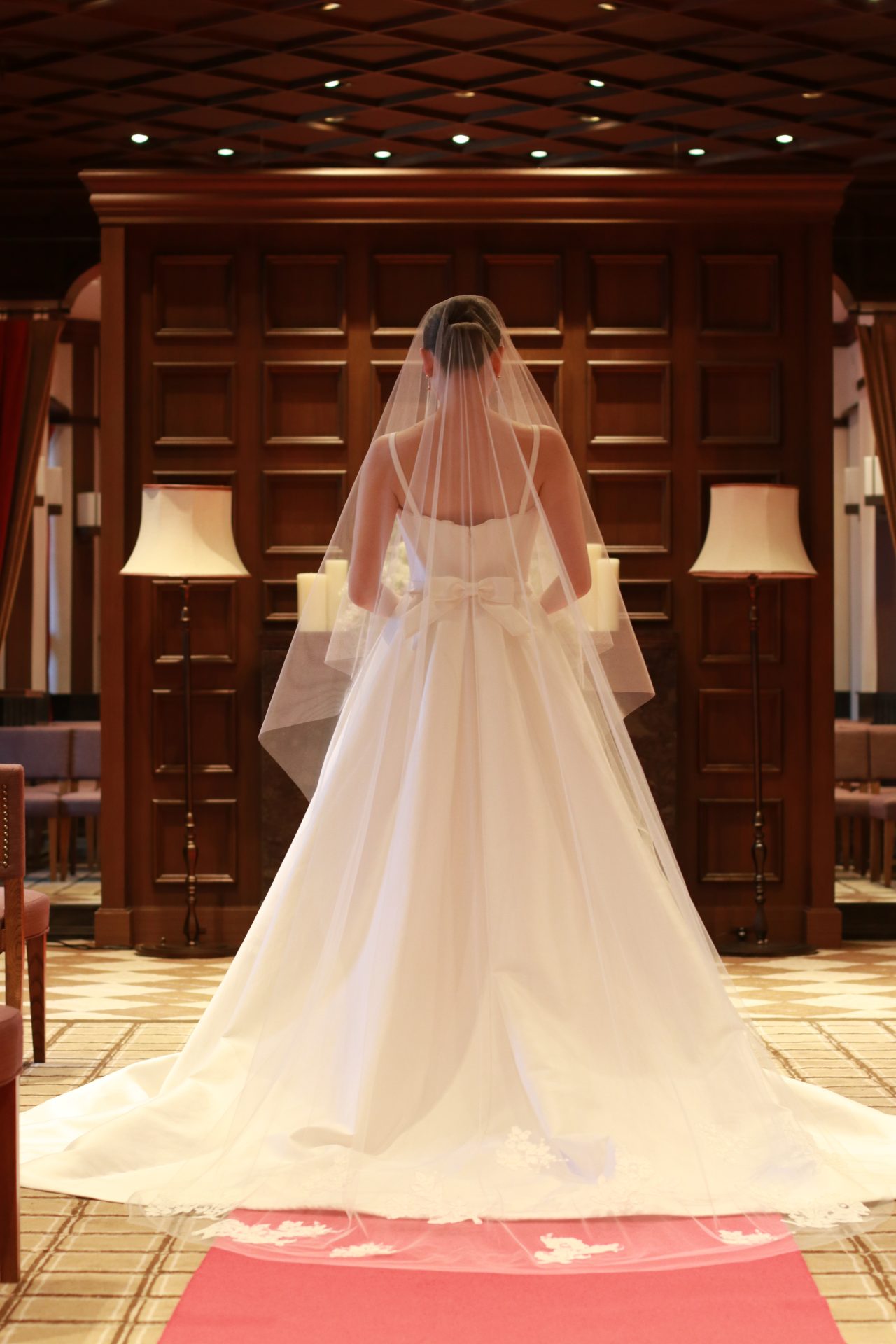 バックスタイルの腰元に施されたキャッチ―なリボンが印象的なシンプルなキャロリーナへレラのウェディングドレスには美しいコードリバーレースの施されたロングベールを合わせることで花を添えます