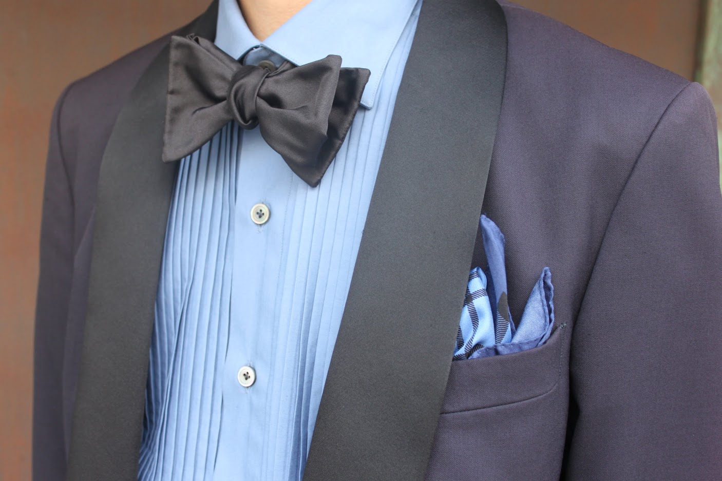 上質なネイビーのフォーマルタキシードのカジュアルダウンスタイル。ブルーのプリーツシャツにブラックのボウタイ。ギンガムチェックのブルーチーフでカジュアルになり過ぎない落ち着いたカジュアルスタイル。