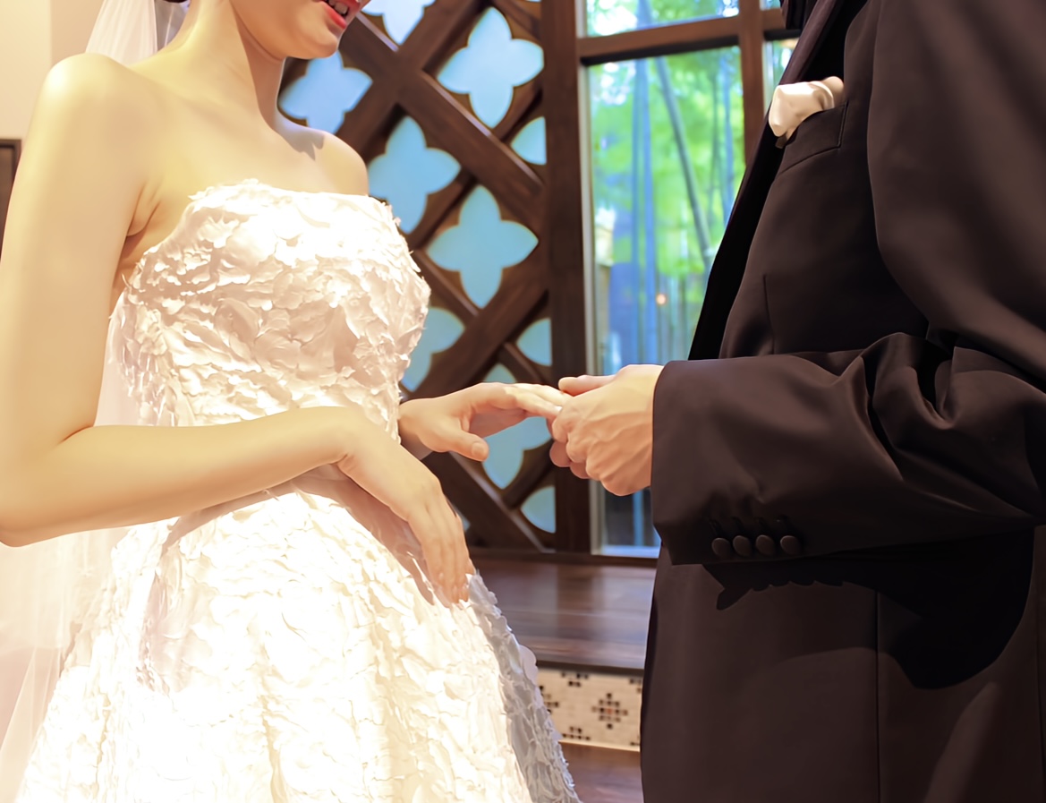 ブラックのタキシードとトリートで取り扱いのあるリームアクラのレーザーカットが綺麗なAラインのウェディングドレスを着用して行うチャペルの中での指輪交換