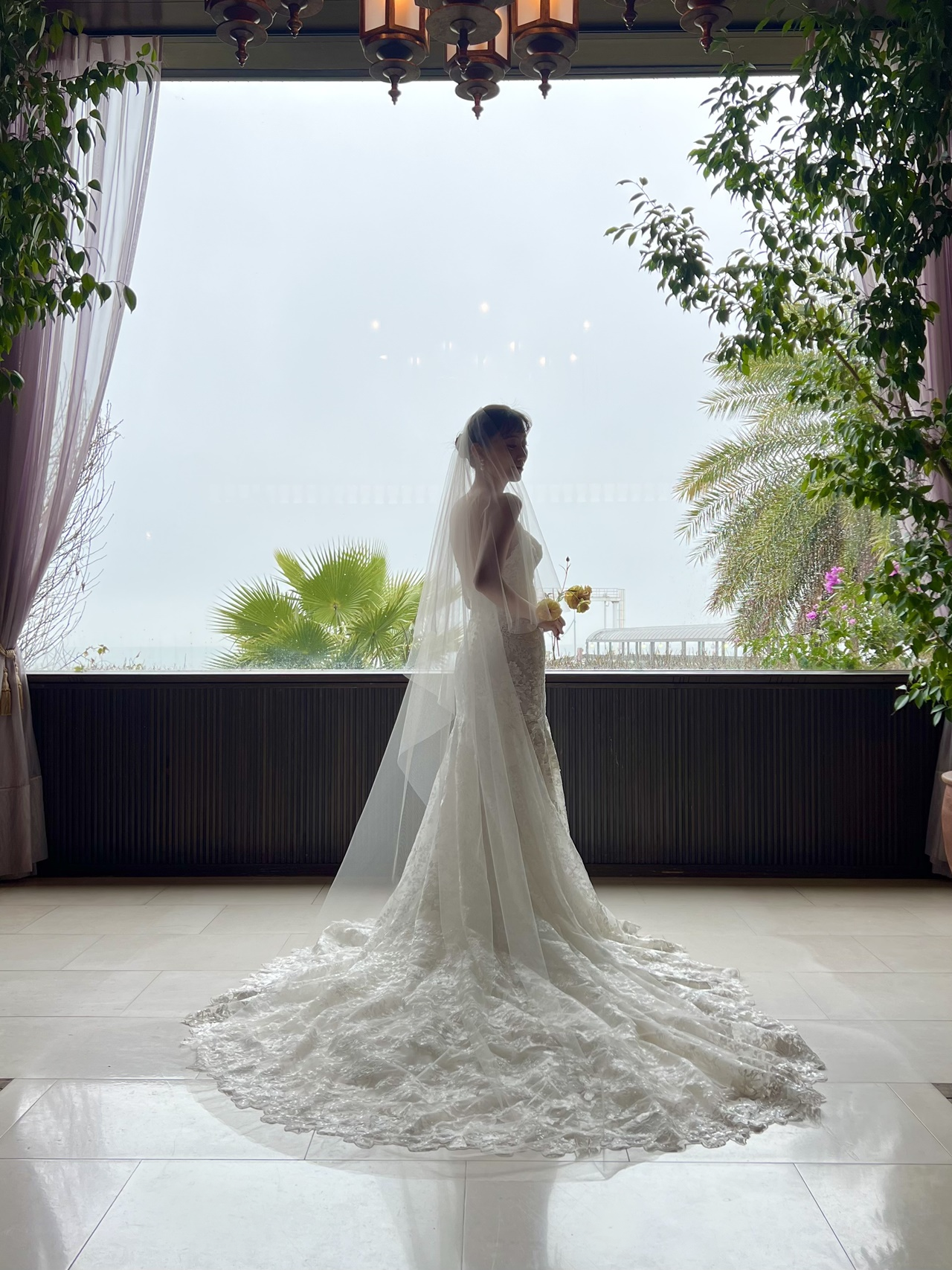 モニーク ルイリエ ブリスが生み出す繊細で優美なマーメイドラインは、花嫁が持つ曲線美をより一層引き立たせます。ヒップラインから大胆に広がるシルエットは、サイズオーダーだからこそ叶えることのできる女性らしいしなやかさを演出し、品格のある雰囲気を纏わせるのです。花嫁の美しさを活かすように、デコルテラインには贅沢に施されたレースの花びらが華やかさをプラス。そして純白ながらも光沢感のある糸で織りなすレースは甘くなりすぎず、どこか凛とした印象で、洗練された花嫁姿を叶えます。陽光が優しく差し込むザ ルイガンズ スパ&リゾートのチャペルでは、散りばめられたクリアなスパンコールは花嫁が動く度にきらめき、圧倒的な存在感を放つことでしょう。決して華美になりすぎず、柔らかな陽光の反射で上品にきらめくスパンコールに、ゲストも思わず魅入ってしまうほど。ヘアスタイルは挙式らしくシンプルにまとめたアップスタイルに、柔らかな透明感をプラスするロングベールを纏いエアリーな質感を。自然体でありながらも、パールのイヤリングをあわせることでクラシカルな気品さを感じさせる優美なコーディネートが完成します。